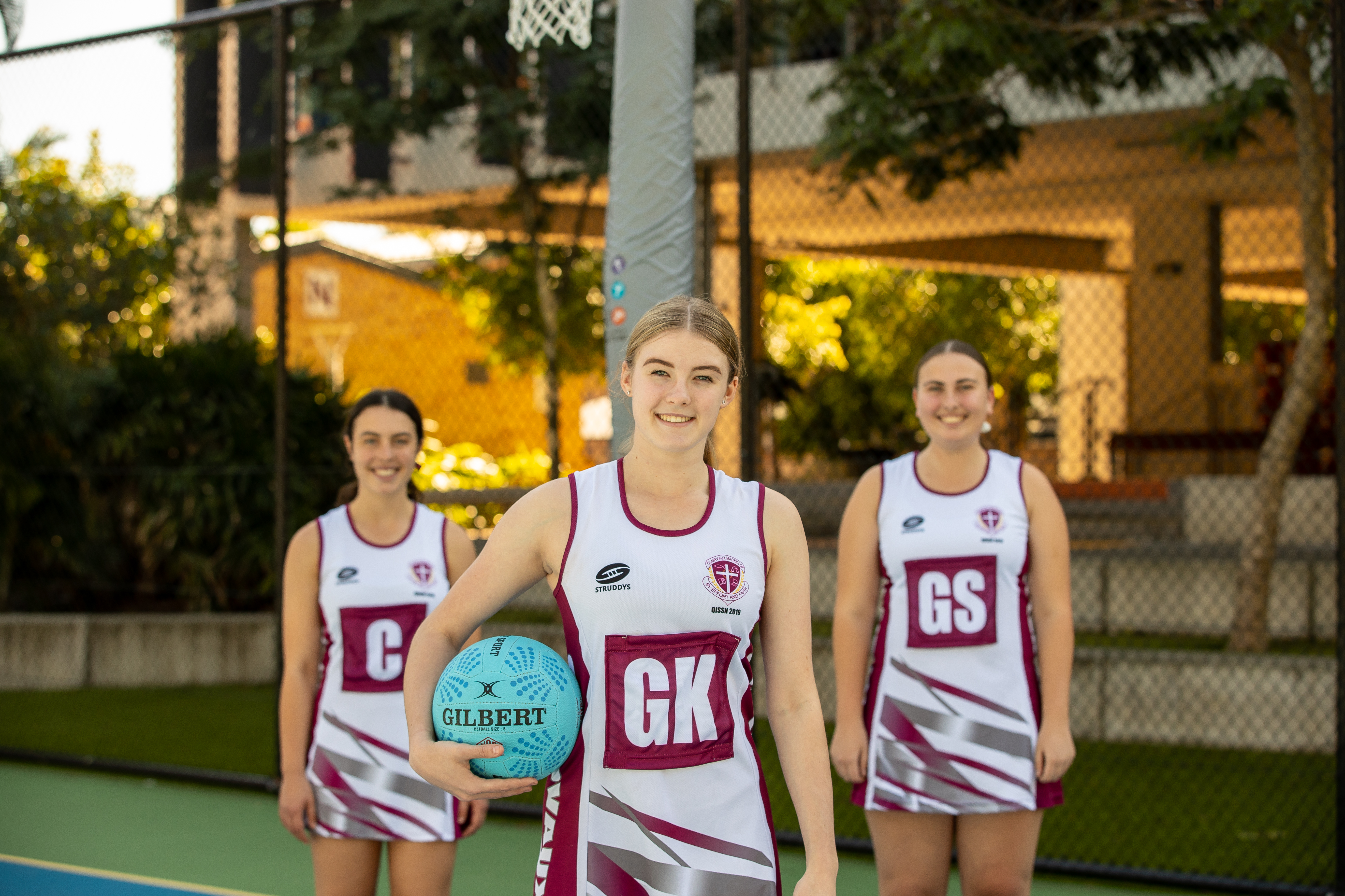 Three students dressed in netball uniforms. One is holding a netball while the other two stand behind her. 
